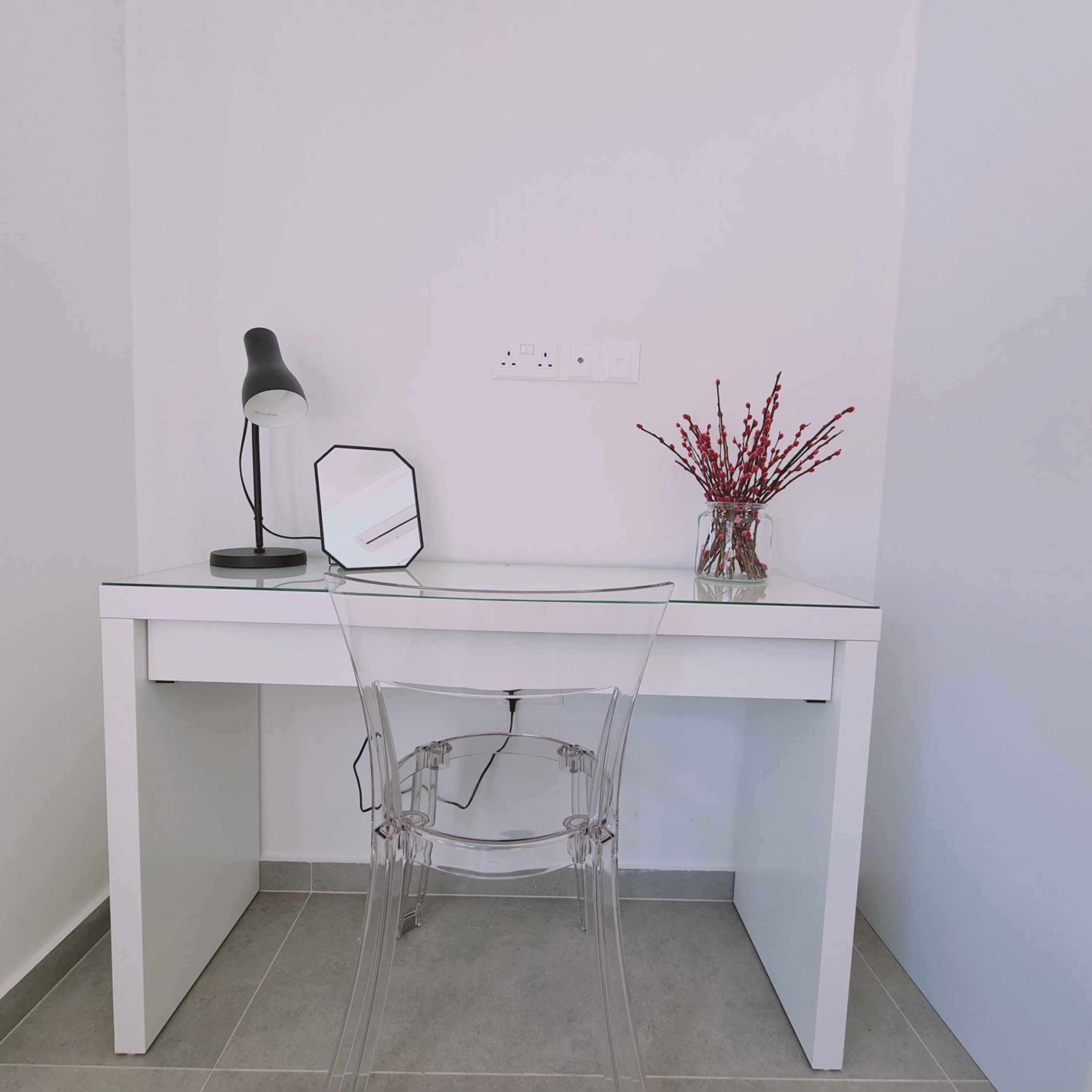 A white desk with a clear chair, a lamp, a mirror, and a vase with red branches.