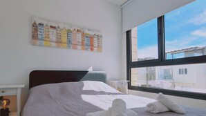 Ein ordentlich bezogenes Bett mit gesteppter Decke und aufgerollten Handtüchern, ein Nachttisch mit Lampe, Wandkunst mit bunten Gebäuden und ein großes Fenster mit Blick auf Gebäude.