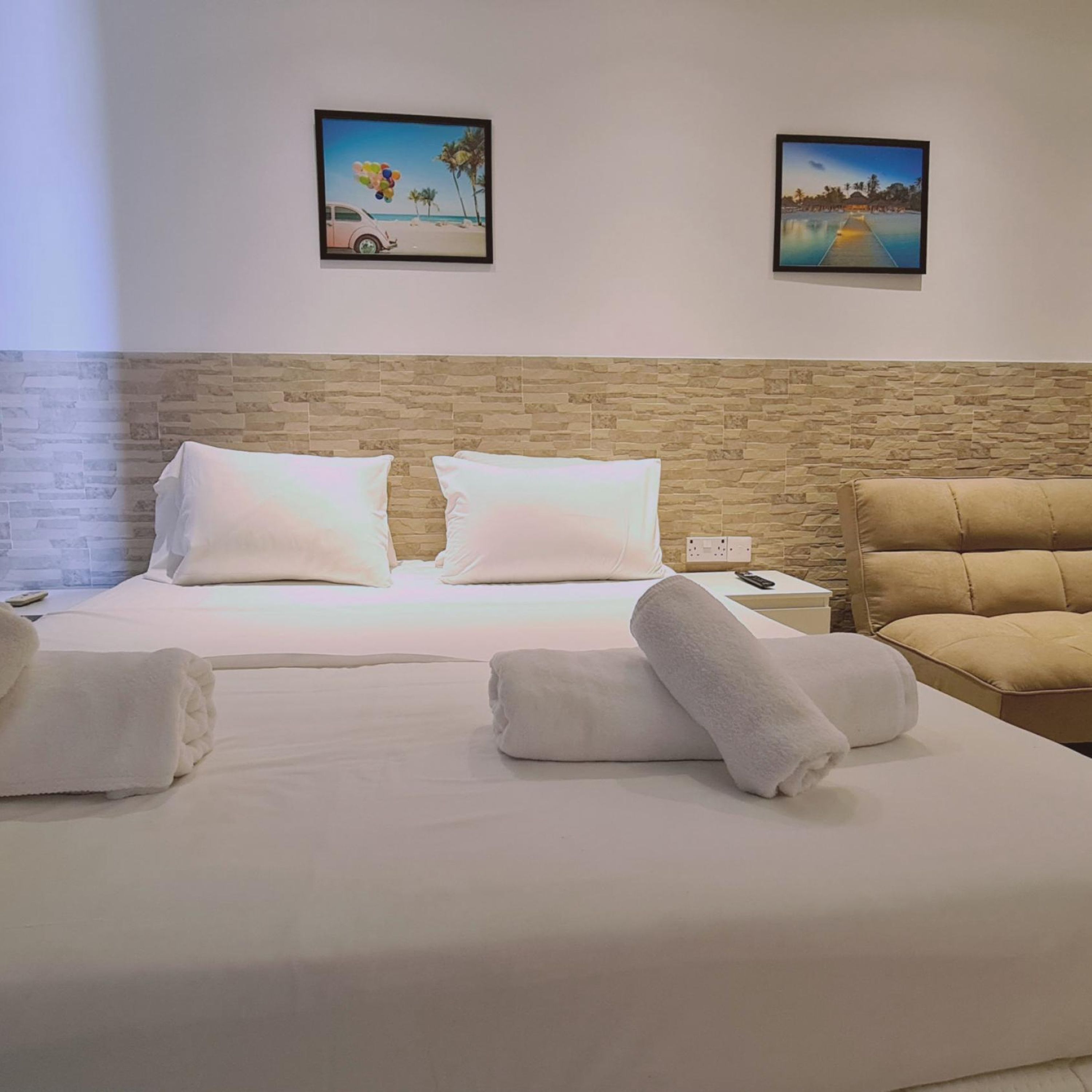 Una habitación de hotel con una cama, dos almohadas, una mesita de noche, un control remoto, una toalla enrollada, una silla y dos cuadros en la pared.