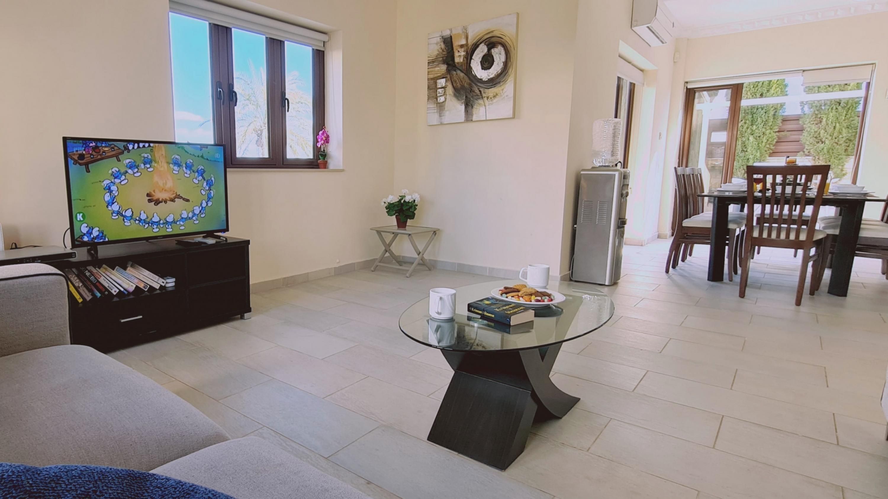 A living room with a flat-screen TV, a glass coffee table with books and a mug, a sofa, a small table with a potted plant, and a dining area with chairs and a table.