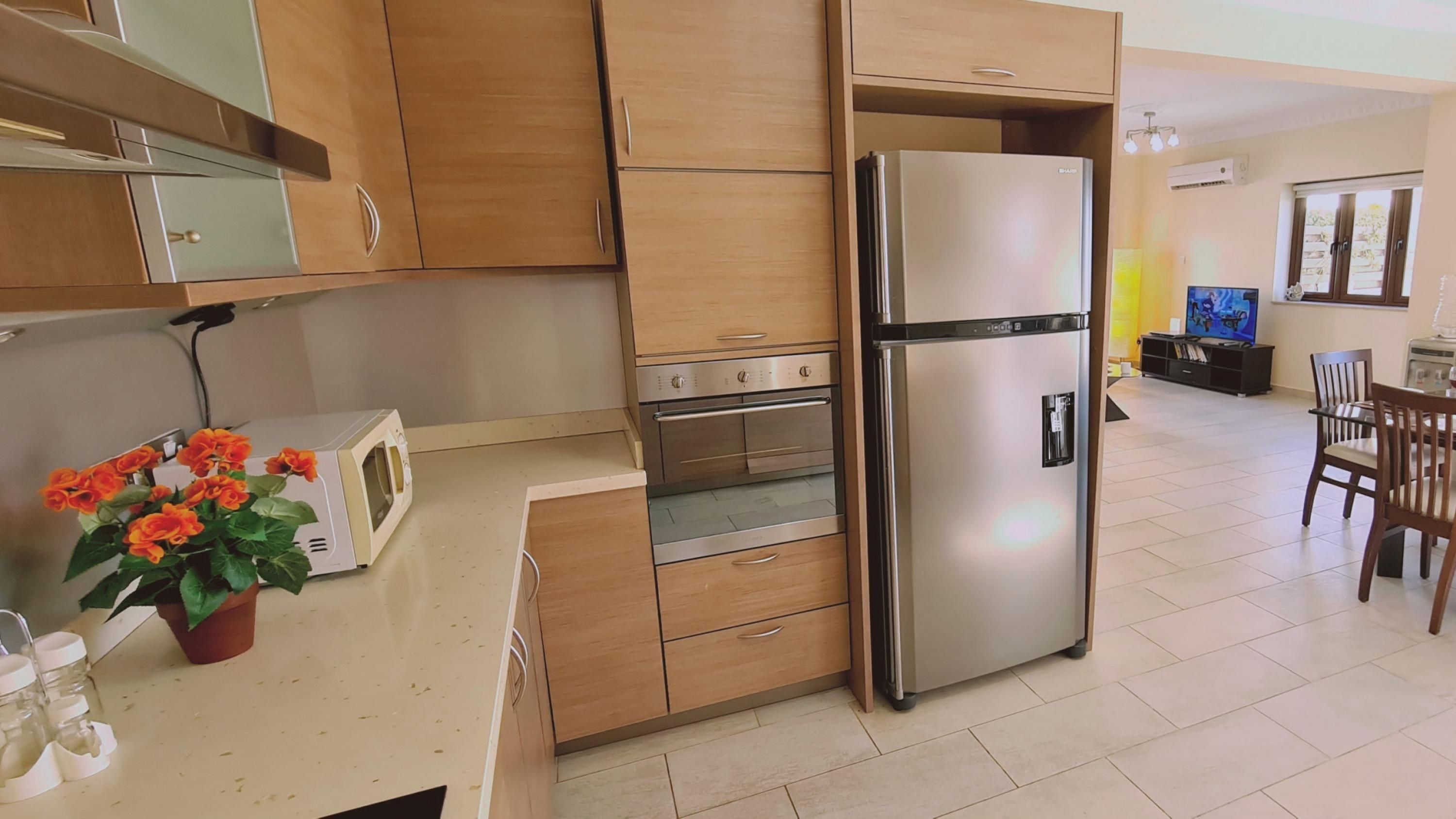 A modern kitchen with wooden cabinets, a stainless steel refrigerator, an oven, and a microwave. There is a dining area with a table and chairs, and a television in the background.