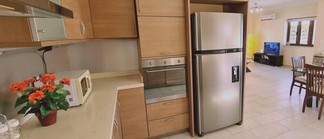 A modern kitchen with wooden cabinets, a stainless steel refrigerator, an oven, and a microwave. There is a dining area with a table and chairs, and a television in the background.