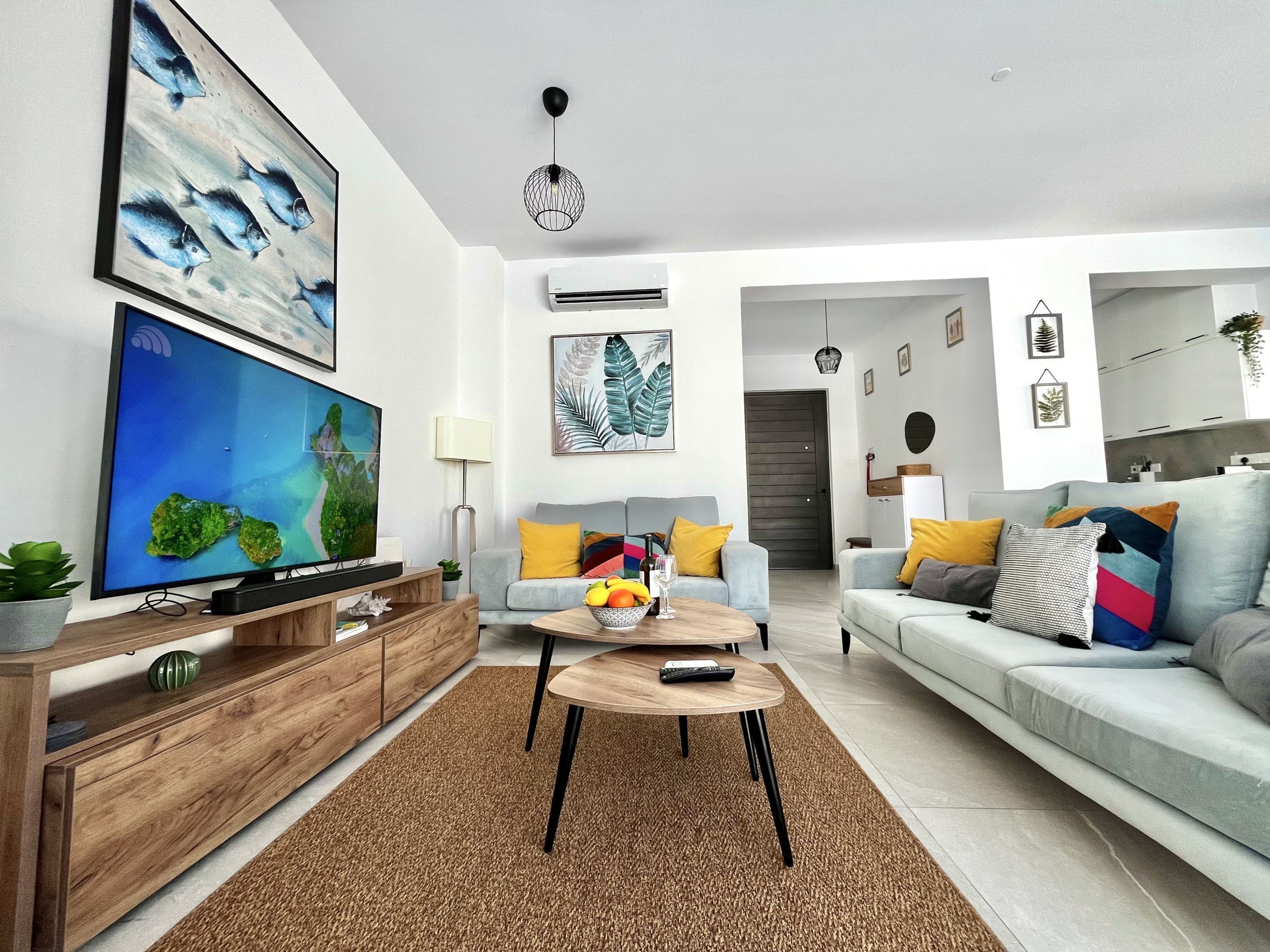Una sala moderna con un televisor de pantalla plana, un mueble bajo de madera, una mesa de centro redonda y dos sofás con cojines de colores.