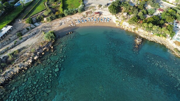 Vista aerea di una spiaggia con acque limpide, un litorale roccioso e una fila di lettini con ombrelloni.