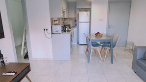 Cozinha moderna com armários brancos, micro-ondas e geladeira. Possui um espaço de jantar com mesa e cadeiras, e um sofá cinza na sala de estar.