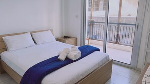 Quarto com cama, mesa de cabeceira e vista para uma varanda através de uma porta de correr de vidro.