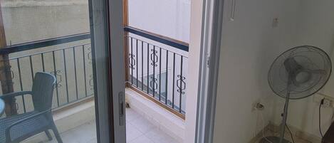 A balcony with a chair and a fan, a window with a view of the sea, and an air conditioning unit on the wall.