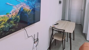 Un salon moderne avec un téléviseur à écran plat fixé au mur, une table à manger en bois avec deux chaises, et une porte visible au fond.