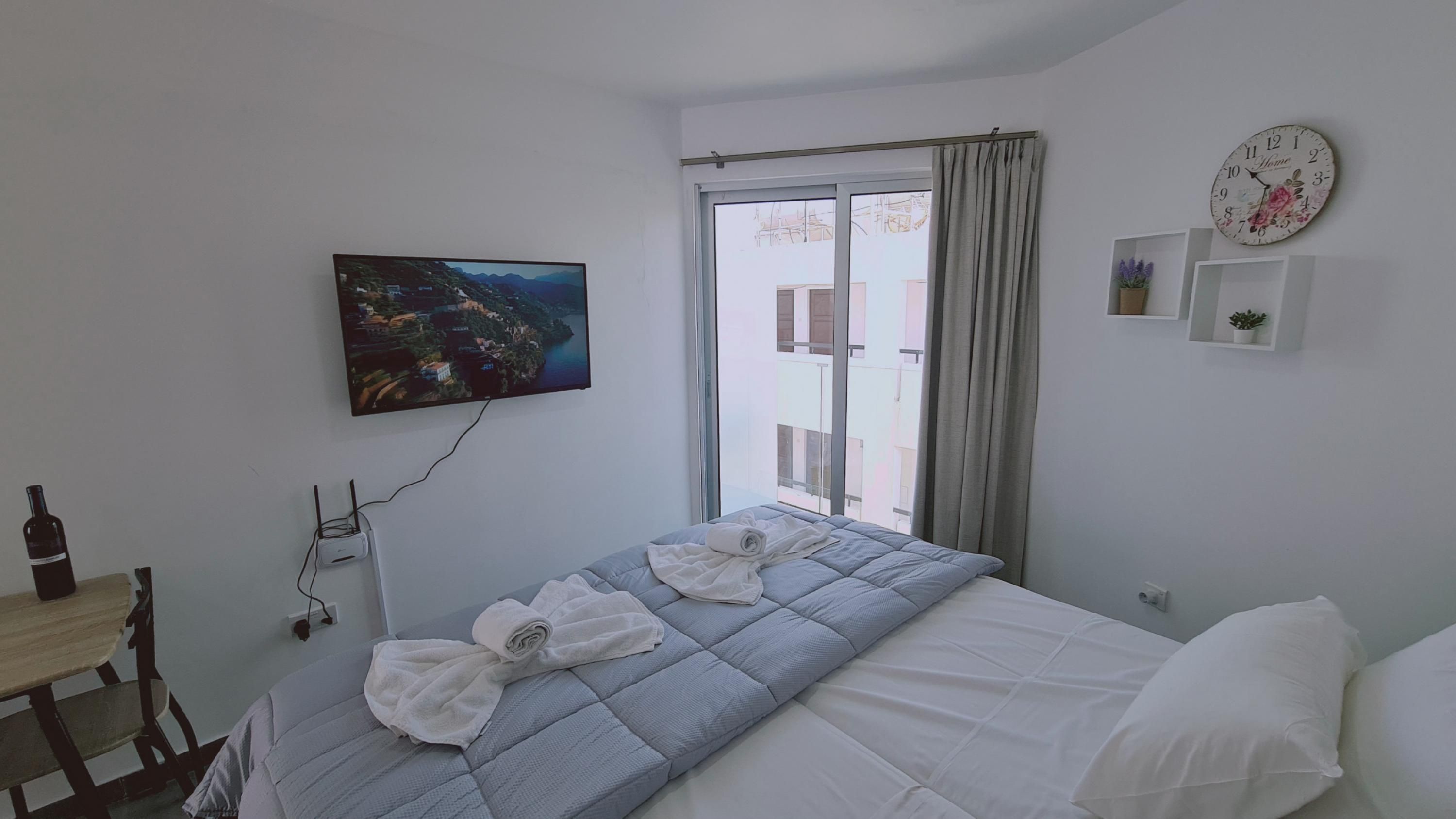 A bedroom with a bed, a TV mounted on the wall, a window with curtains, and a small shelf with a clock and a plant.