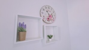 Une horloge murale ornée de motifs floraux, sur laquelle figure le mot « Home ». Il y a deux étagères blanches, l’une avec une plante en pot et l’autre avec une plante plus petite.