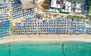 An aerial view of a beach resort with rows of blue umbrellas, a sandy beach, and clear turquoise water. - 100 AN - Napa Living 18 (Ayia Napa)