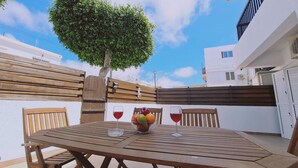 Ein Holztisch mit Obstschale und zwei Weingläsern, umringt von Holzstühlen, vor einer weißen Wand mit Baum und Holzzaun.