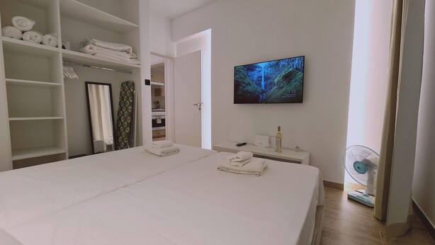Ein Hotelzimmer mit einem großen Bett, einem an der Wand befestigten Fernseher, einem kleinen Nachttisch mit einer Flasche und Fernbedienung sowie einem Ventilator in der Nähe des Fensters.