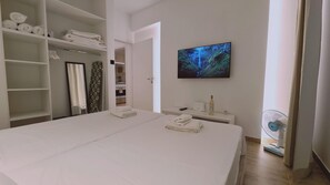 A hotel room with a large bed, a television mounted on the wall, a small bedside table with a bottle and remote, and a fan near the window. - 72 AN - Stay Napa Velvet (Ayia Napa)