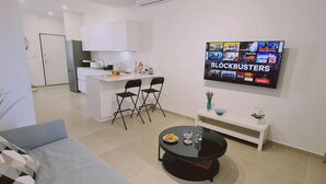 A modern living room with a grey sofa, a black coffee table, and a TV displaying a movie selection. A kitchen with white cabinets and a dining area with two black chairs are visible in the background. - 46 PRO - Stay Anemone 20 (Paralimni)