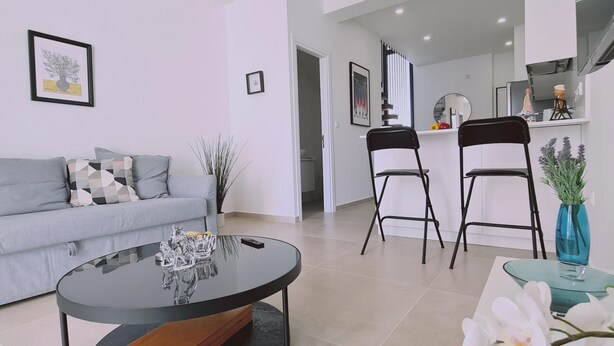 A modern living room with a grey sofa, a coffee table, and two black chairs. There is a kitchen area in the background with white cabinets and a microwave. - 46 PRO - Stay Anemone 20 (Paralimni)
