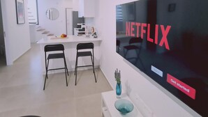 A modern living space with a large flat-screen TV displaying 'NETFLIX' and a 'Get started' button. There are two black chairs, a white console table with a blue vase, and a kitchen area in the background. - 46 PRO - Stay Anemone 20 (Paralimni)