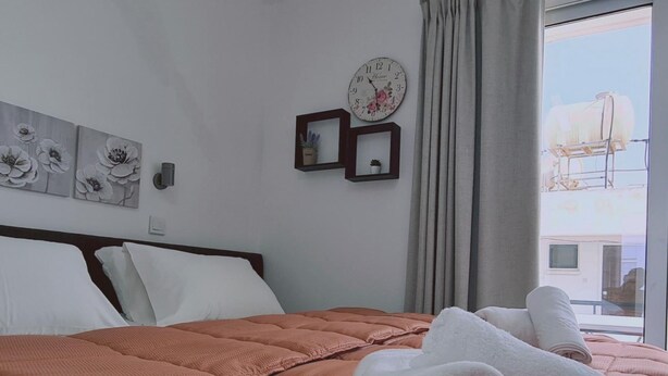 Un letto rifatto con cura, con una coperta rosa, cuscini bianchi e asciugamani bianchi arrotolati su una parete bianca con un orologio e quadri incorniciati.