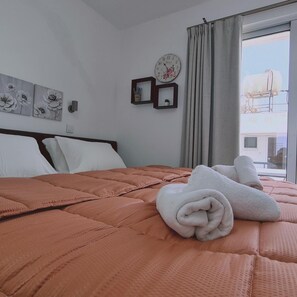 A neatly made bed with a pink quilt, white pillows, and rolled white towels on a white wall with a clock and framed pictures. - 100 AN - Napa Living 21 (Ayia Napa)