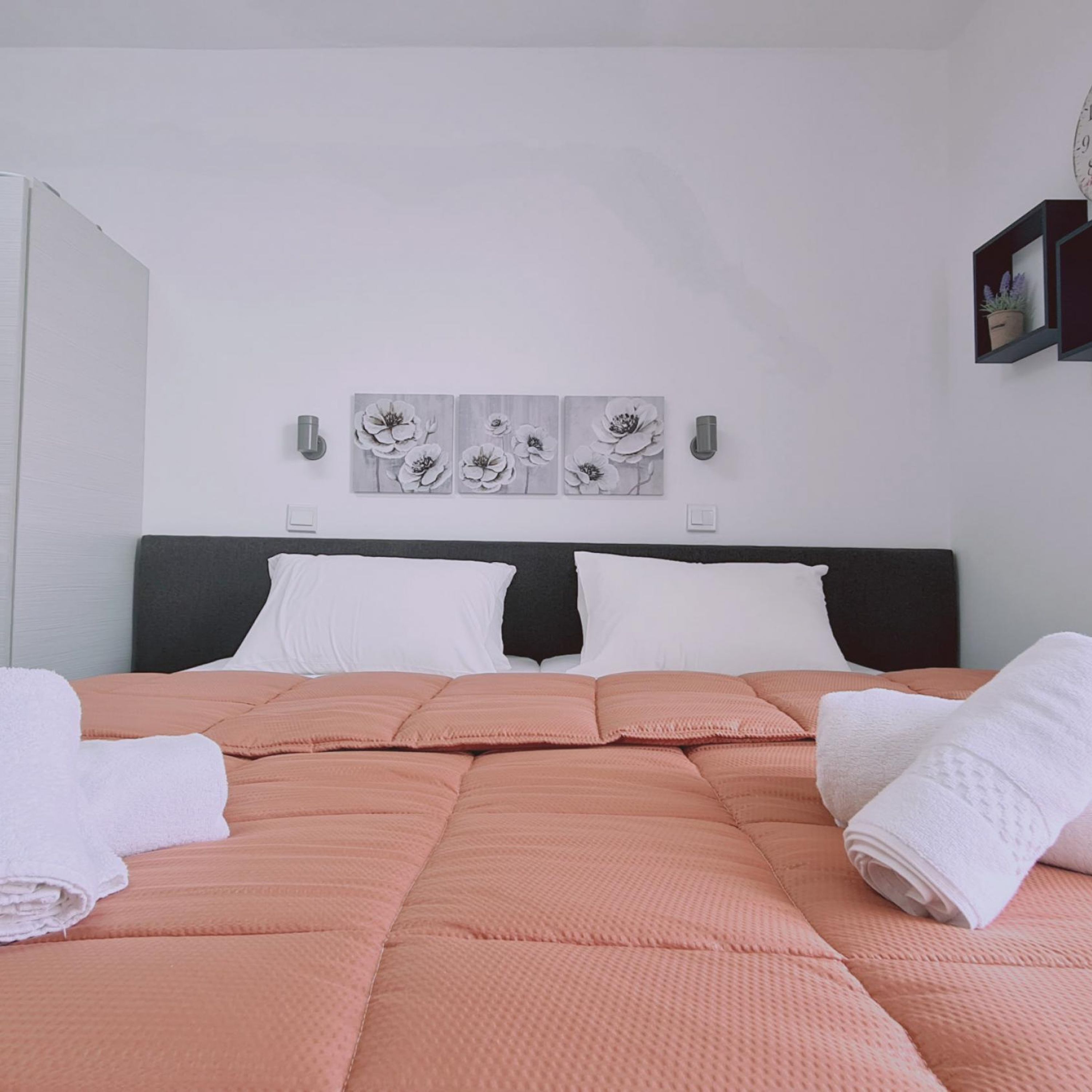 Una cama bien hecha con colcha rosa, almohadas blancas y una toalla blanca a un lado. Encima de la cama, tres cuadros de flores colgadas en la pared.