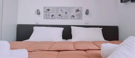 Una cama bien hecha con colcha rosa, almohadas blancas y una toalla blanca a un lado. Encima de la cama, tres cuadros de flores colgadas en la pared.