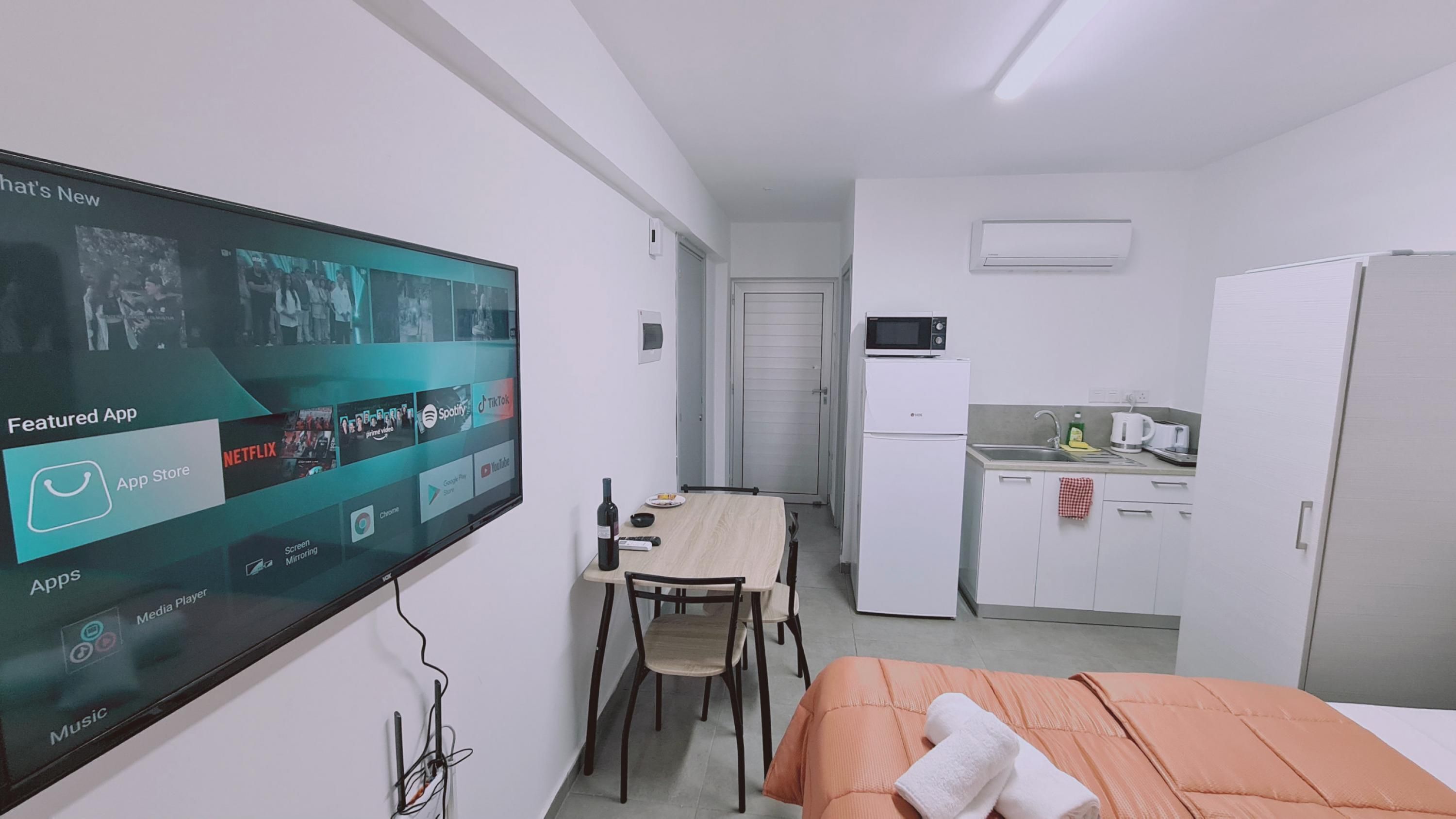 A compact hotel room with a bed, a TV displaying a streaming service interface, a small kitchenette, and a dining area.