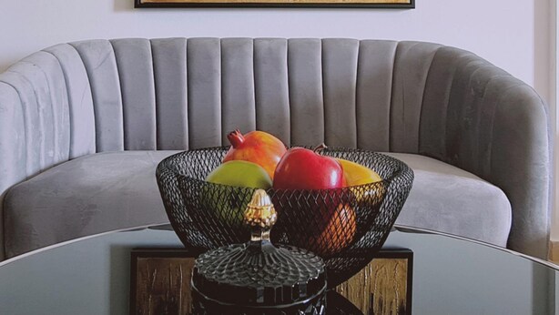 Ein graues Sofa mit einem schwarz gerahmten Gemälde eines Waldes. Davor ein Glastisch mit einem schwarzen Korb voller Früchte.