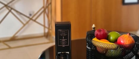 Un dispositivo electrónico negro sobre una mesa de cristal con una cesta de frutas que contiene manzanas y naranjas.