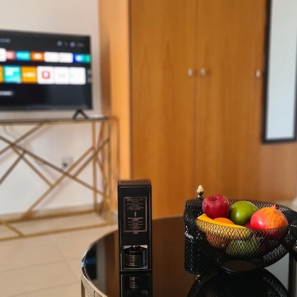 Musta elektroninen laite lasipöydällä, pöydällä on myös hedelmäkori, jossa on omenoita ja appelsiineja.