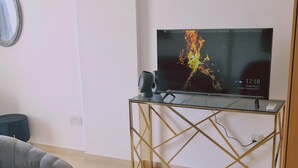 Ein Wohnzimmer mit einem Flachbildfernseher auf einem goldfarbenen Ständer, einem grauen Sofa und einem Couchtisch mit einer Dekoration.