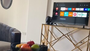 Ein Wohnzimmer mit einem an der Wand befestigten Flachbildfernseher, einem runden Glastisch mit Früchten und einer Fernbedienung sowie einem modernen Nachttisch mit goldener Rahmen.