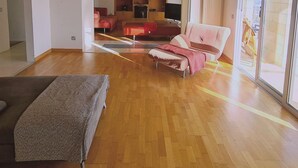 A spacious living room with a wooden floor, a sofa, a red armchair, and a television. - 95 1 TLA - Stay Sunset Grand Palazzo (Tala)