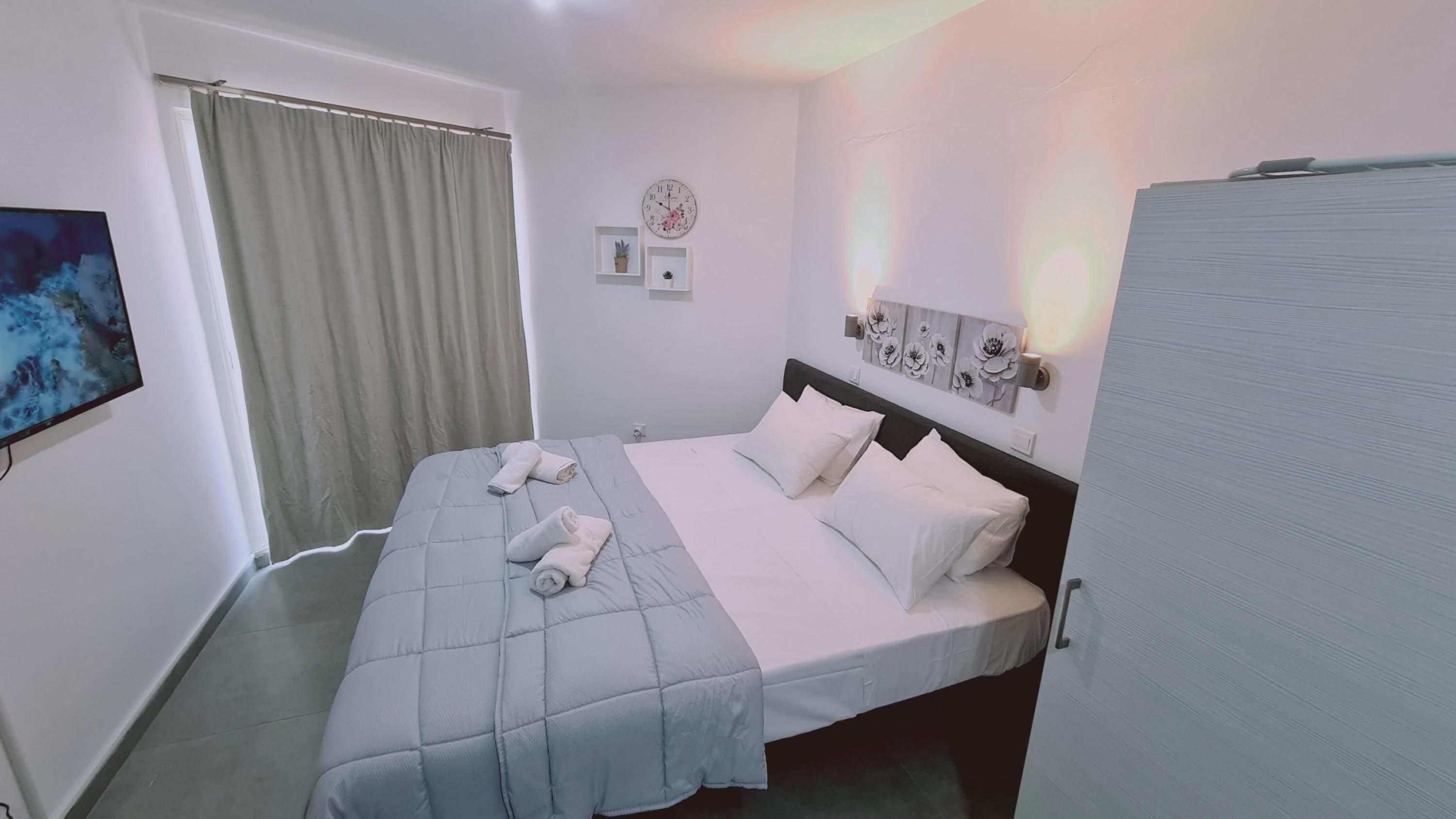 A hotel room with a large bed, a flat-screen TV, a clock, and a window with curtains.