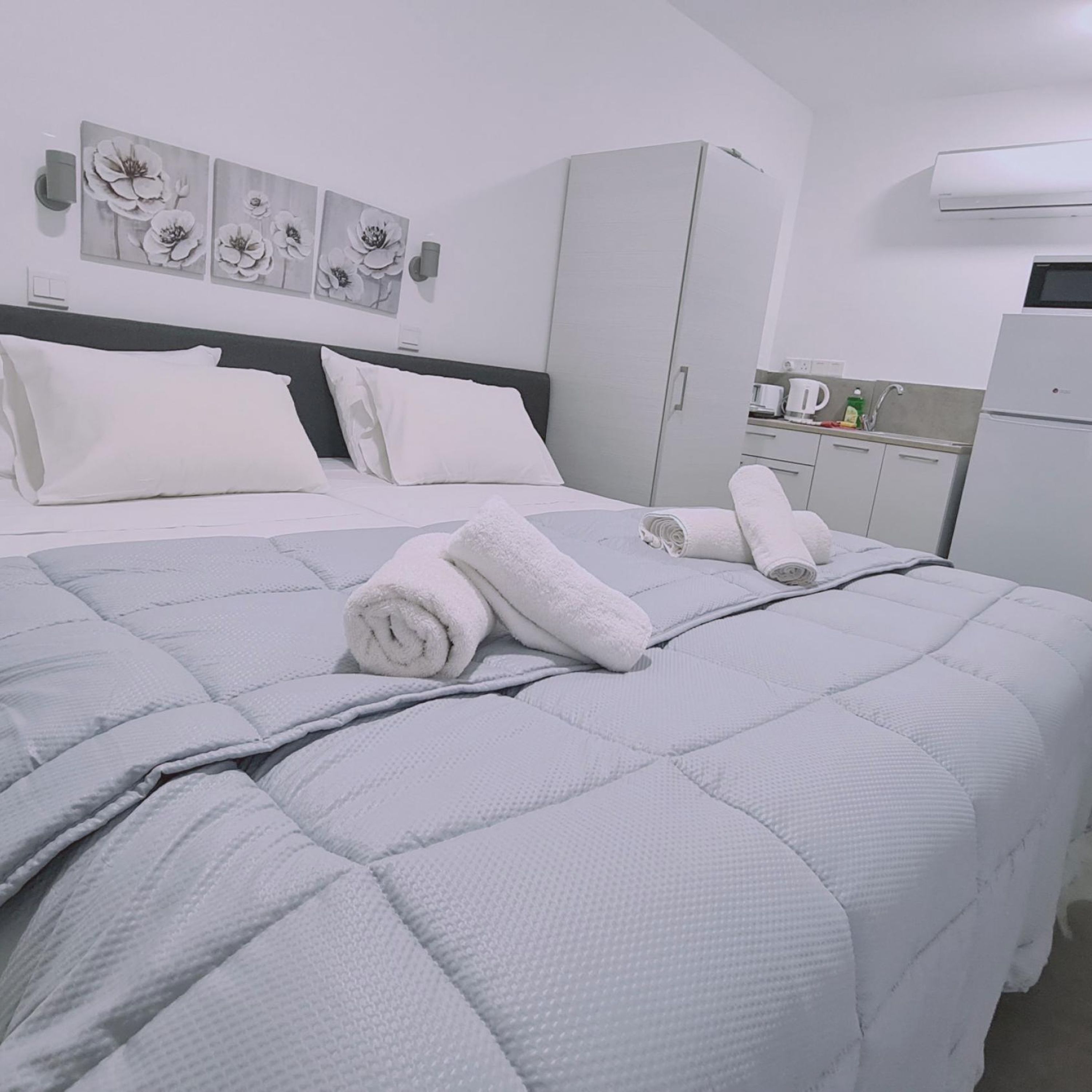 Een net opgemaakt bed met een grijs gebreid dekbed, witte kussens en opgerolde witte handdoeken. Er is een grijze garderobe, een kleine kitchenette met een koffiezetapparaat en een airconditioning op de muur.