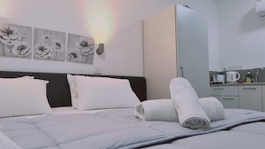 A neatly made bed with white linens and rolled towels, a wall-mounted light fixture, and a small kitchenette area with a kettle and coffee maker.