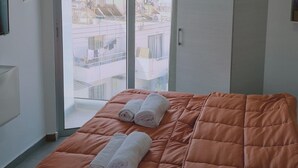 Ein Schlafzimmer mit Bett, Kleiderschrank und Blick auf Gebäude durch ein großes Fenster.