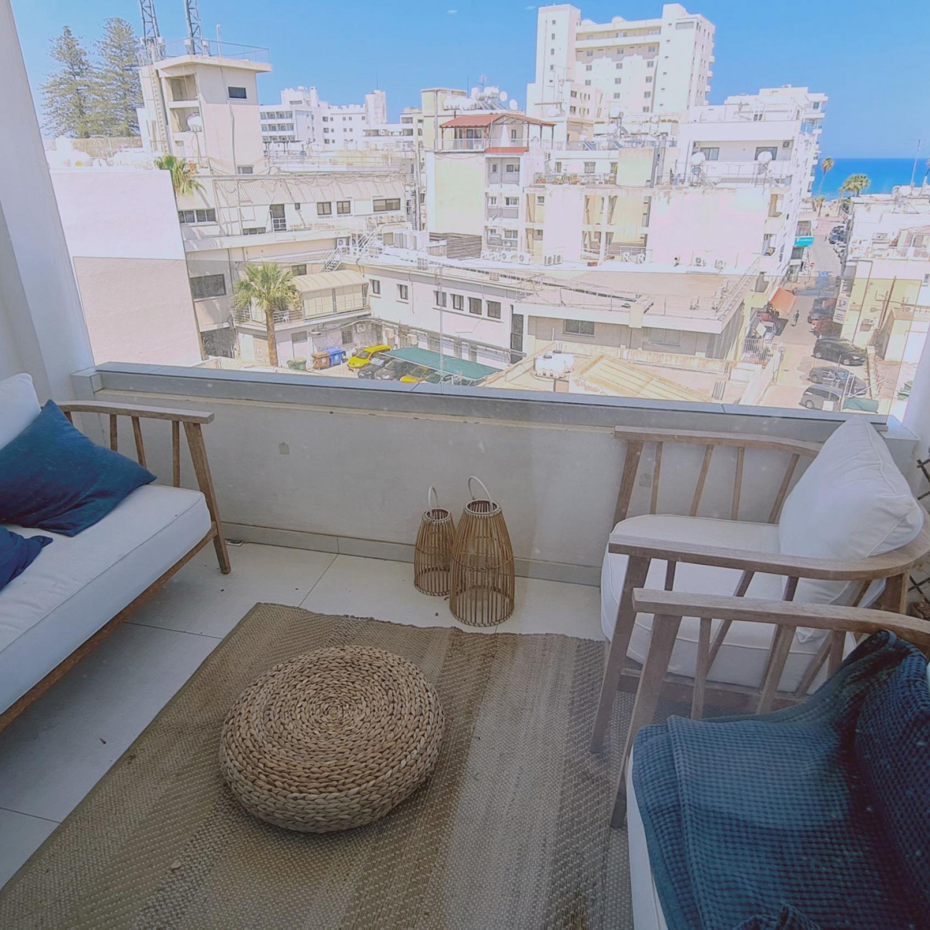 A balcony with a view of a cityscape, a sofa, a chair, and a woven pouf.