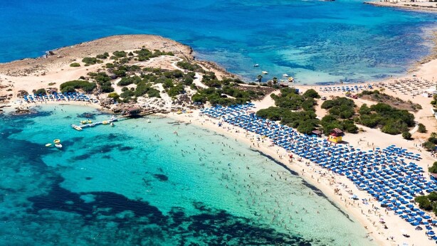 An aerial view of a beach with clear turquoise waters, a sandy shore, and numerous blue umbrellas.