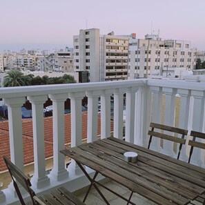 Een balkon met witte hekwerkleuningen, een houten tafel en een metalen beker. Op de achtergrond zien we meerlaags gebouwen met balkons.