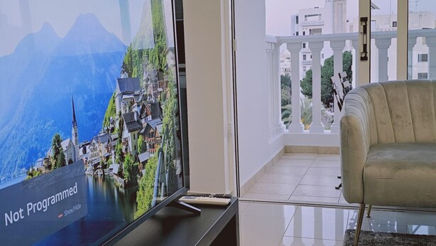A modern living room with a flat-screen TV displaying a scenic mountain village. - 104 NIC - 403 Stay Cosmo City Suite (Nicosia)