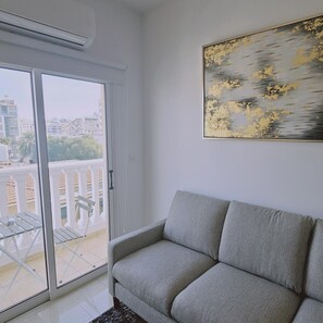 Un soggiorno con un divano grigio, una porta scorrevole in vetro che dà su un balcone esterno e un quadro appeso al muro.
