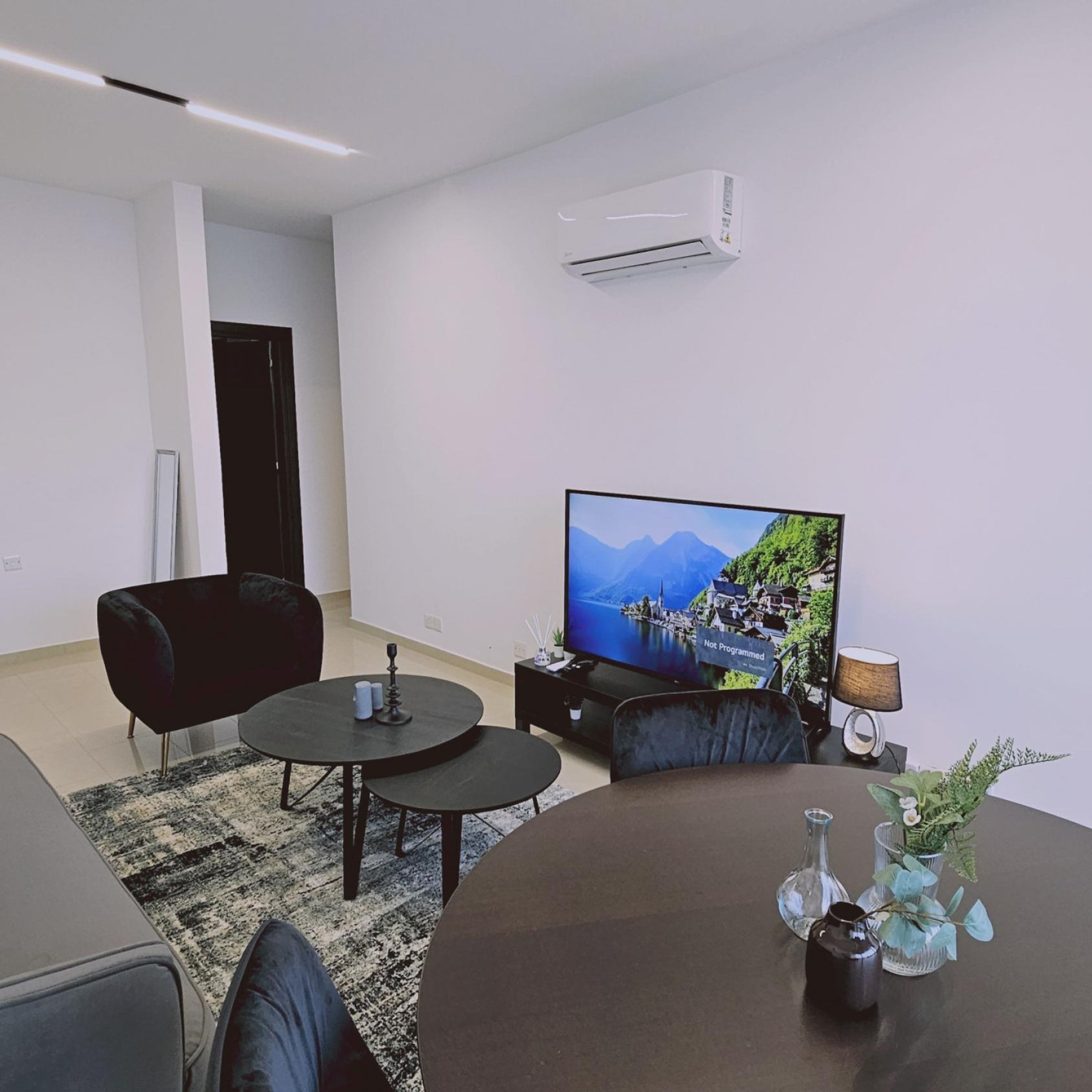 Ein modernes Wohnzimmer mit einem Flachbildfernseher, einem schwarzen Sofa, einem Couchtisch und einem kleinen Beistelltisch mit Lampe und Dekorationsgegenständen.