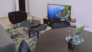 Un soggiorno moderno con una televisione a schermo piatto, un divano nero, un tavolino da caffè e un piccolo tavolino laterale con una lampada e oggetti decorativi.
