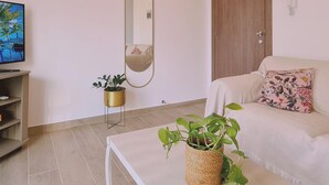 A living room with a white sofa, a coffee table with a plant, a TV on a stand, a wooden door, and a large mirror on the wall. - 83 Lim3 - Stay Sunlit Apartment (Limassol)