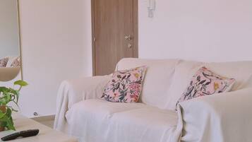 A living room with a beige sofa, two floral cushions, a remote control, and a small table with a plant.