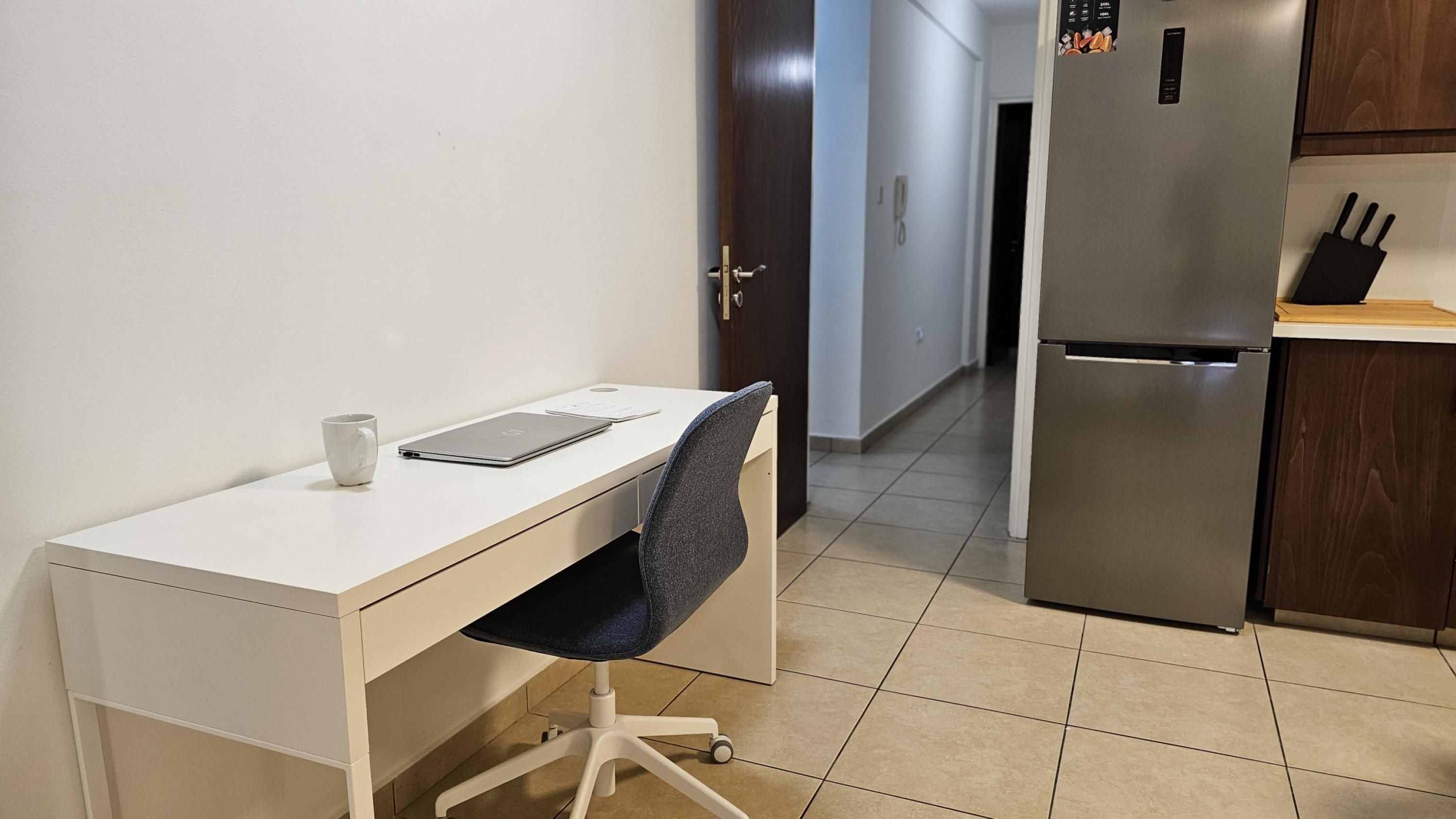 Una habitación pequeña y limpia con un escritorio blanco, una silla, una computadora portátil y una taza. Se ve una puerta que da a otra habitación y un refrigerador al fondo.