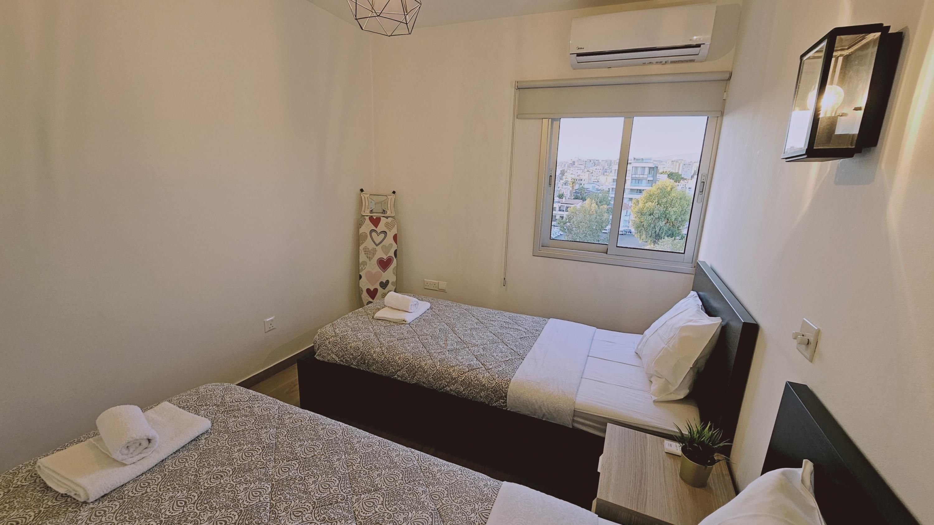 Une petite chambre propre, avec deux lits, une fenêtre donnant sur des immeubles, un climatiseur et un luminaire fixé au mur.