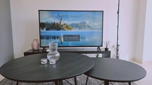 Un televisor de pantalla plana sobre un soporte, una mesa de madera oscura con dos sillas, un jarrón rosa y una pequeña planta.