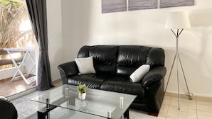 Een moderne woonkamer met een zwarte leren bank, een glazen salontafel met een plant, en een vloerlamp. Boven de bank hangen drie ingekaderde foto’s.