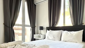 Hotelkamer met een groot bed, grijze gordijnen, nachtkastje met een lamp en uitzicht op buiten.